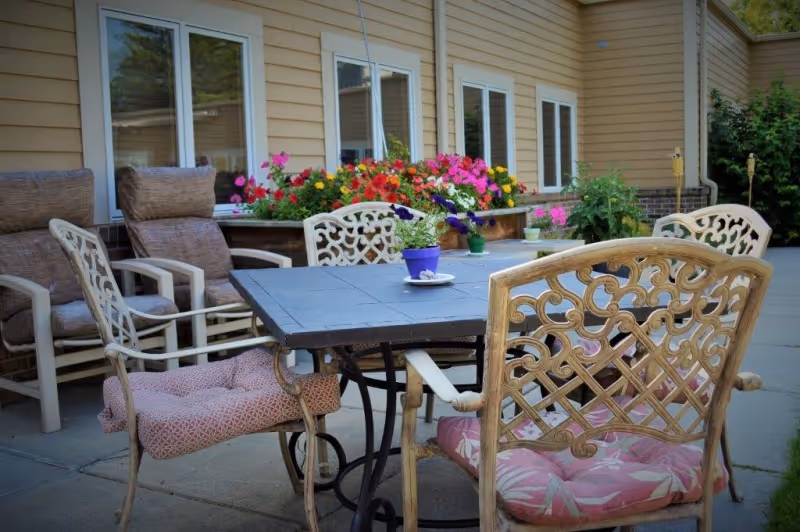 Outdoor patio area with a black metal table surrounded by cushioned chairs, including ornate metal chairs with pink cushions and beige armchairs. Behind the seating area, there is a flower bed with colorful blooming flowers and a building with beige siding and multiple windows.