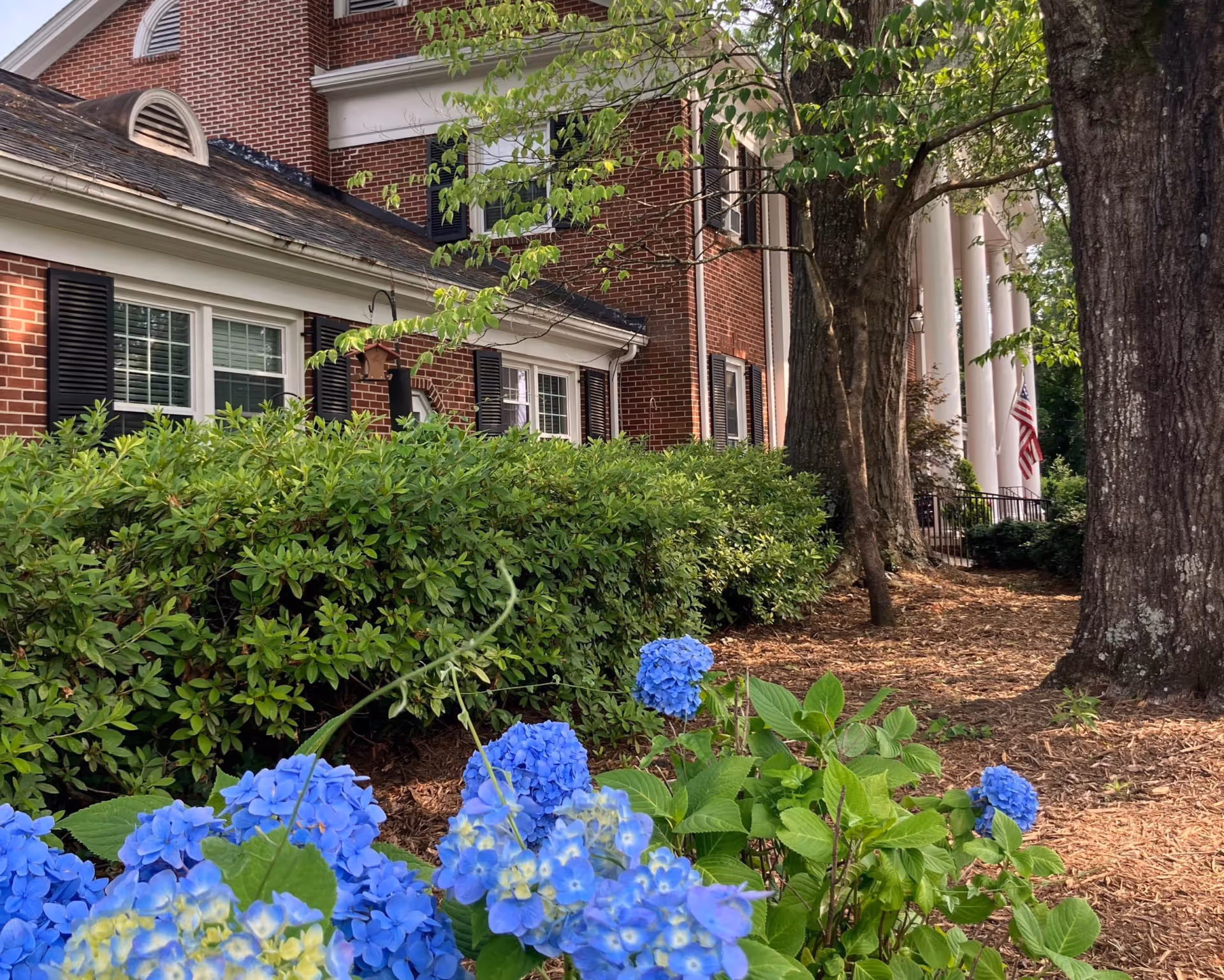 Exterior view of Sarah Gossett Retirement Home with a brick facade, white columns, green shrubs, and blue hydrangeas in the foreground.