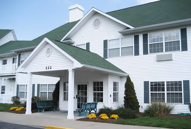 Exterior view of a two-story white building with green roof and green shutters, featuring a covered entrance with benches and landscaping including bushes and yellow flowers.