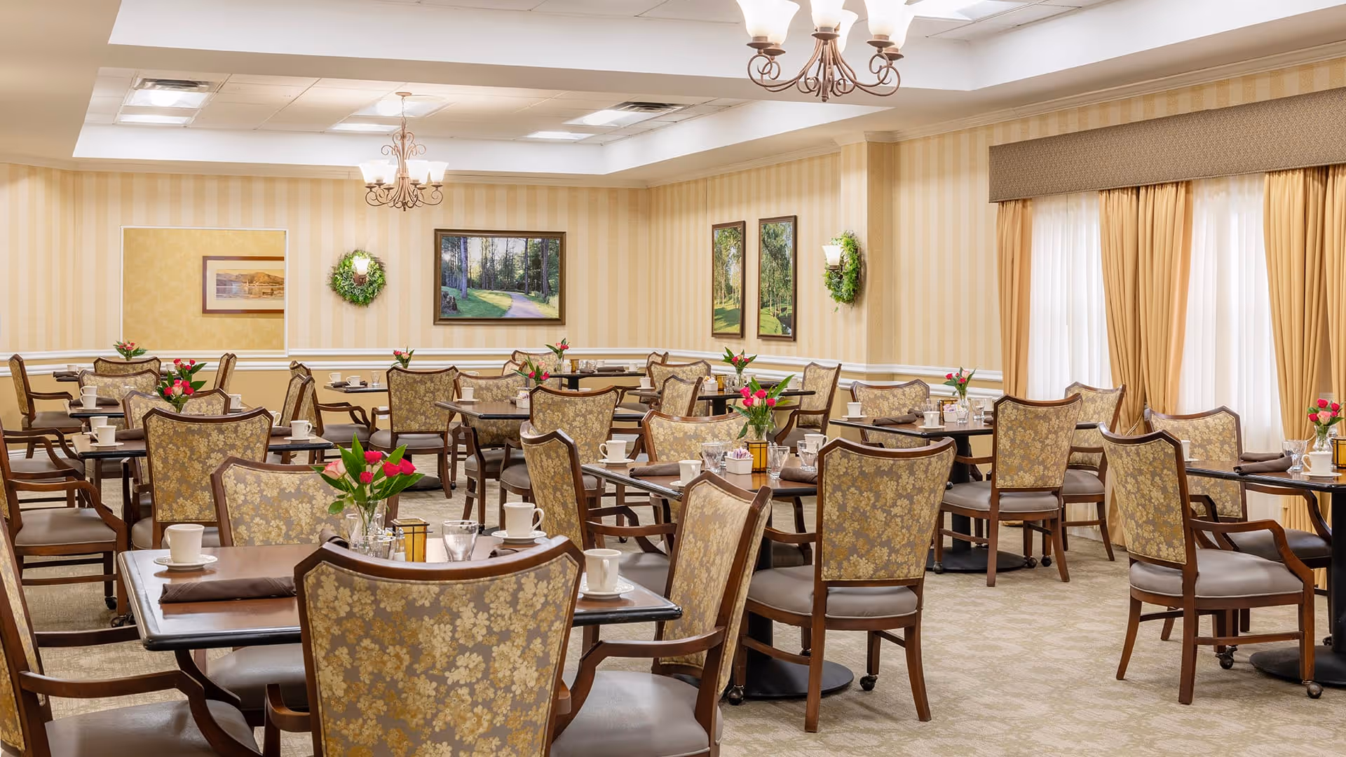 A well-lit dining room with multiple tables and chairs arranged neatly. Each table has a small flower arrangement and place settings including cups and napkins. The room features beige striped wallpaper, framed landscape paintings, wall sconces with greenery, and large windows with beige curtains allowing natural light to enter.