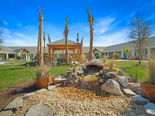Outdoor courtyard area with a small rock waterfall feature surrounded by gravel and plants, palm trees, a wooden gazebo, and a single-story building with a covered walkway in the background under a blue sky.
