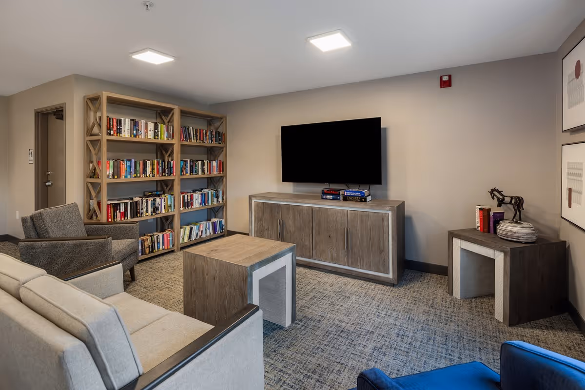 A cozy living room area in a senior living community featuring a light gray sofa, a gray armchair, a wooden coffee table, a wooden TV stand with a flat-screen TV mounted above it, and a wooden bookshelf filled with books. The room has neutral-colored walls, carpeted flooring, and framed artwork on the wall.