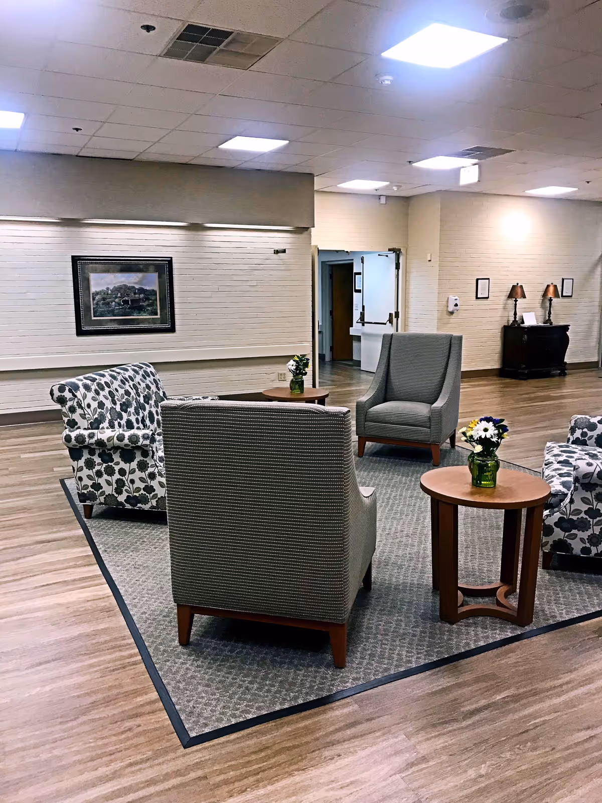 A seating area in a senior living facility with four armchairs arranged around two small wooden tables with flower vases. The room has wood flooring, a carpet under the chairs, a framed picture on the wall, and soft lighting from ceiling fixtures.