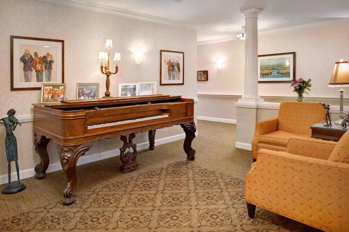 A cozy interior room featuring a vintage wooden piano with framed photos and artwork on the wall above it. To the right, there are two orange upholstered armchairs with a side table holding a lamp and a vase with flowers. The room has patterned carpet, soft wall lighting, and decorative columns.