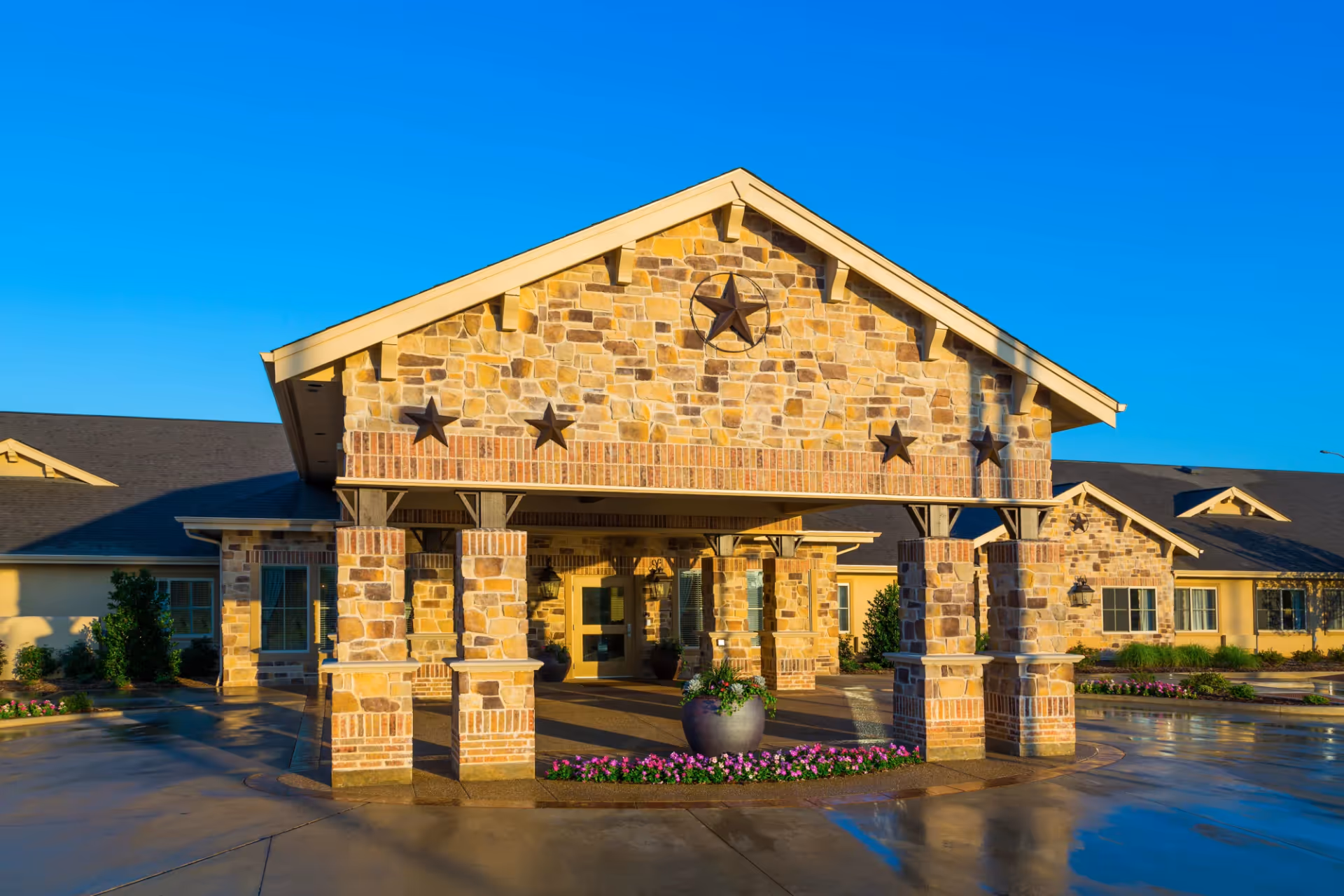 Front exterior view of a senior living facility with a stone and brick facade, featuring a covered entrance supported by stone pillars. The building has decorative stars on the front gable and is surrounded by landscaped flower beds and shrubs under a clear blue sky.