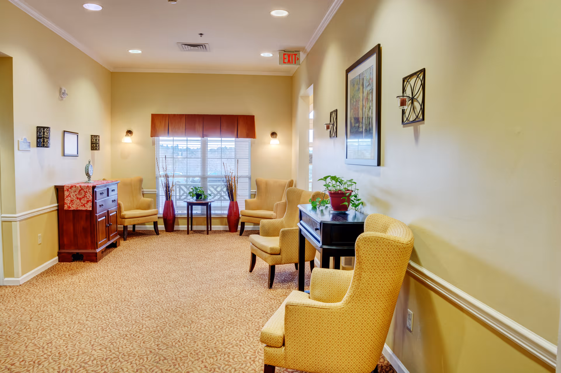 A well-lit sitting area in a senior living facility with beige walls and carpet. The space features several cushioned armchairs arranged along the walls, a small wooden table with a potted plant, and a wooden cabinet with decorative items. Wall sconces and framed artwork decorate the walls, and a window with a valance lets in natural light. An exit sign is visible above a doorway.