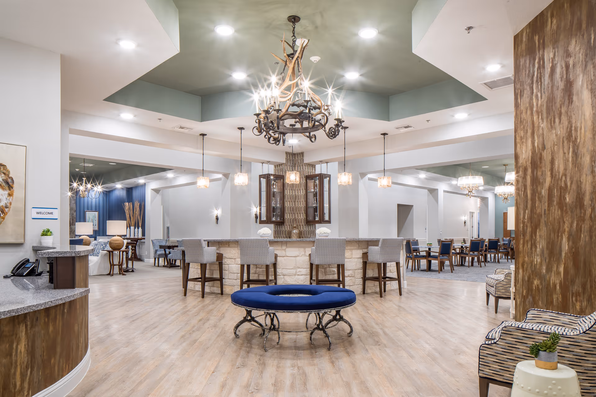 Spacious senior living facility common area with a central chandelier, round blue bench, bar counter and dining tables.