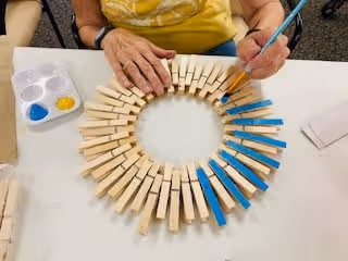 A person painting wooden clothespins blue, which are clipped around a circular frame, on a white table. A small paint palette with blue and yellow paint is visible on the left side of the table.
