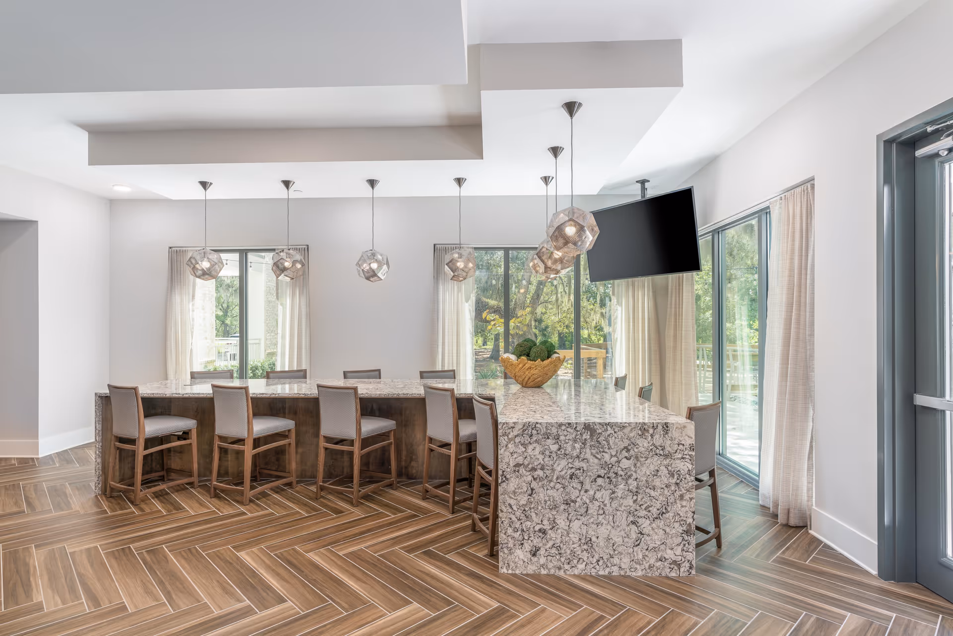 A modern dining area with a large marble island countertop surrounded by wooden chairs with gray cushions. Above the island, there are six geometric pendant lights hanging from the ceiling. Large windows with sheer curtains allow natural light to fill the room, and a flat-screen TV is mounted on the ceiling near the windows. The floor features a wood-patterned herringbone design.