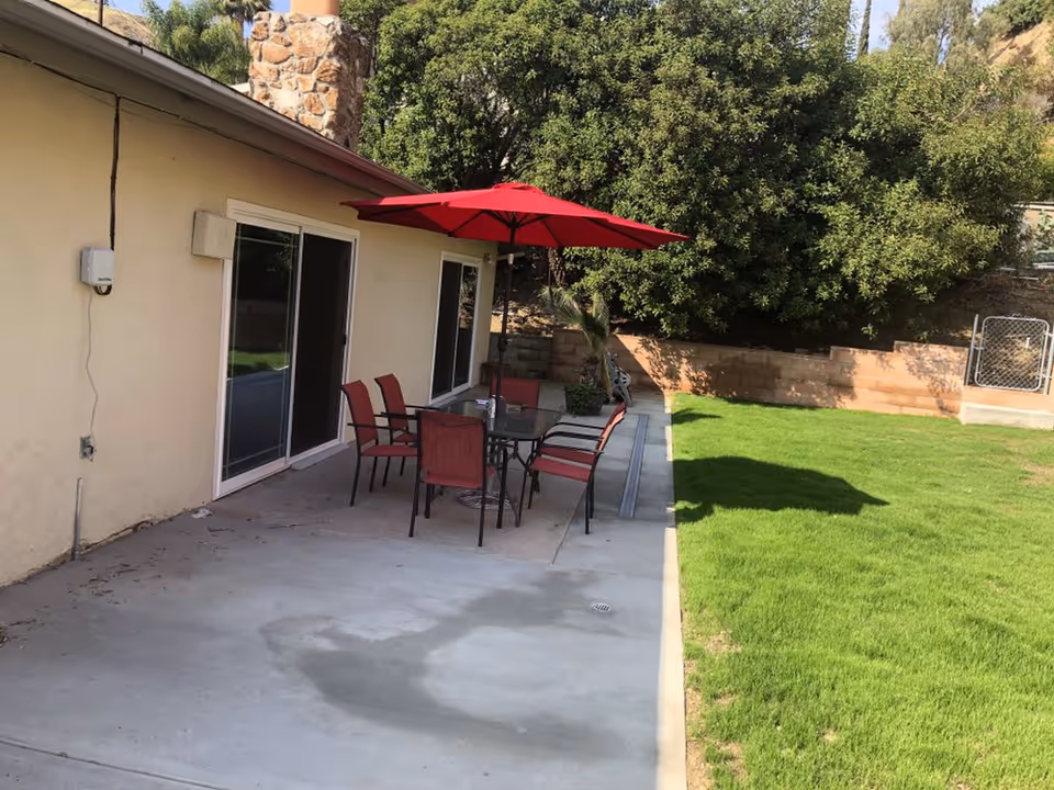 Outdoor patio area with a concrete floor adjacent to a beige building. There is a glass-top table with six red chairs around it and a large red umbrella providing shade. The patio is next to a well-maintained green lawn and bordered by a low brick wall with dense green bushes behind it.