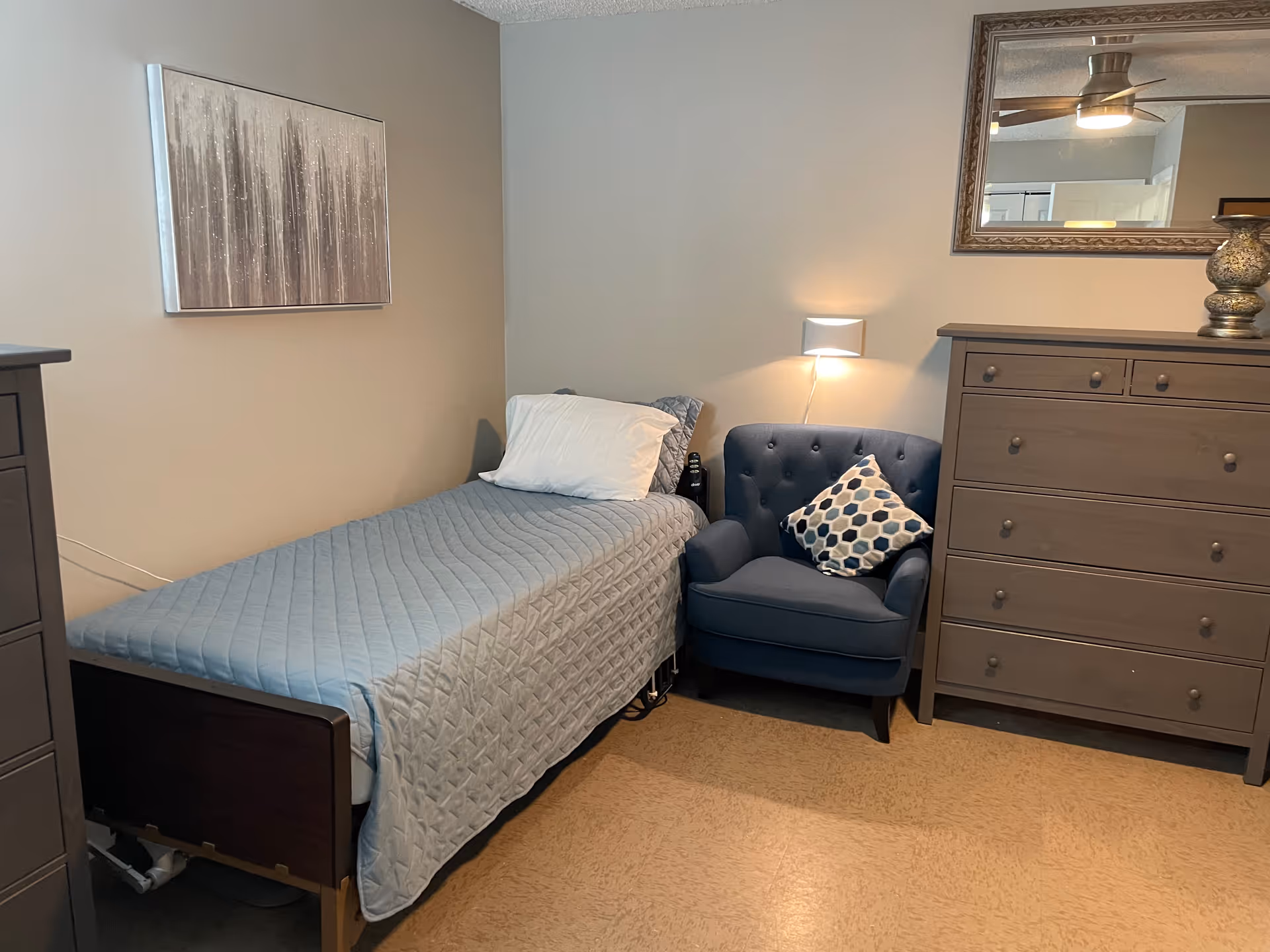 A tidy single bedroom with a bed covered in a blue quilt, a blue armchair with a patterned pillow, and a wooden dresser under a mirror.