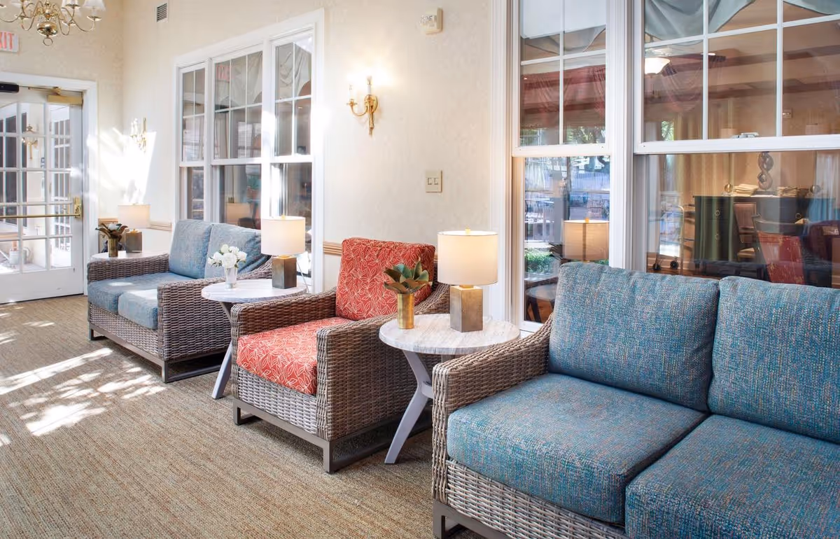 A bright and cozy sitting area with wicker furniture including two blue cushioned sofas and one red cushioned armchair. Each seating piece is paired with a small round table holding a lamp and a small plant. Large windows and glass doors allow natural light to fill the room, creating a welcoming atmosphere.