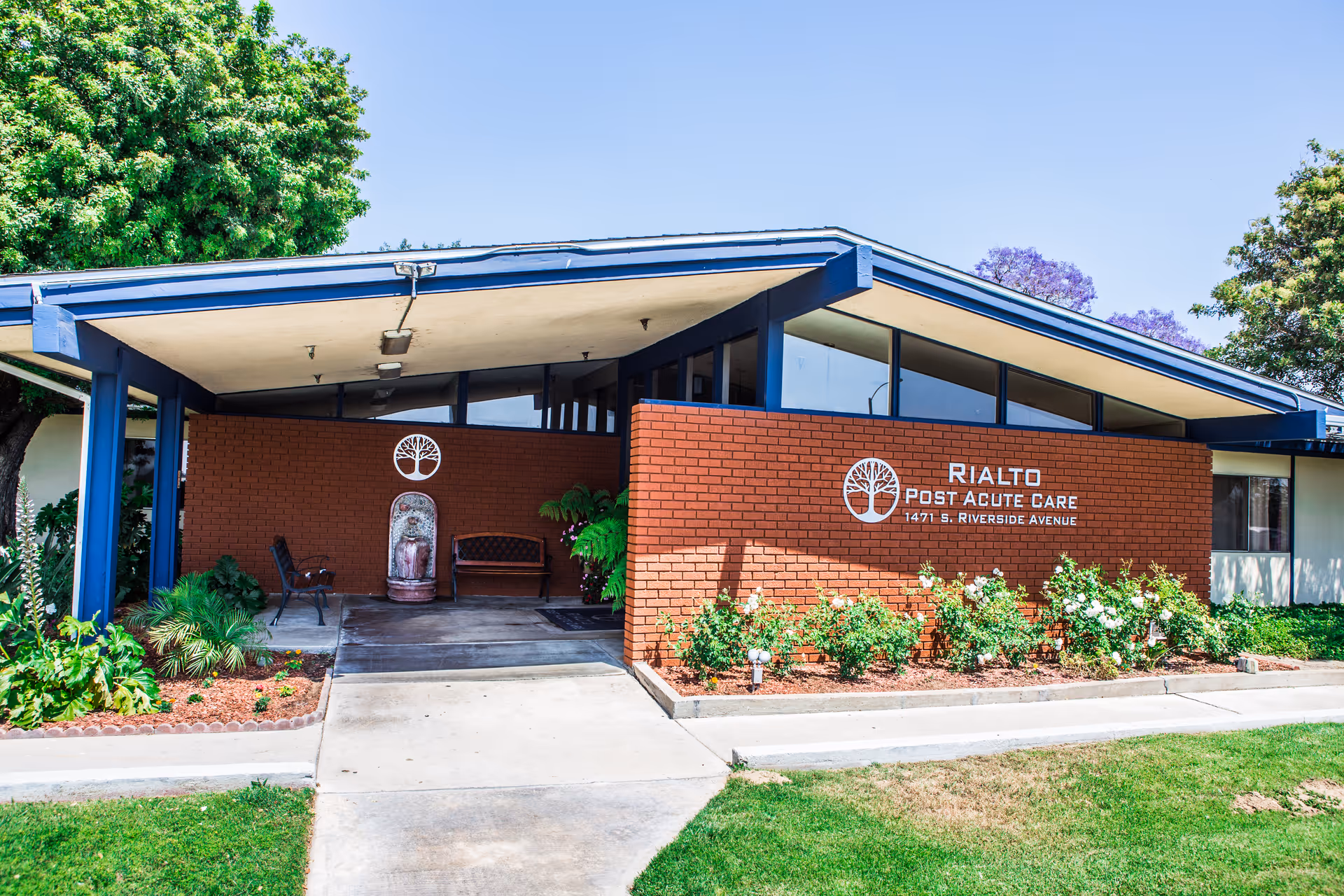 Exterior view of Rialto Post Acute Care building with a covered entrance, brick walls, blue roof supports, and surrounding greenery including bushes and trees.