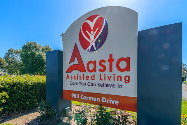 Outdoor sign for Aasta Assisted Living located at 903 Carmen Drive, with a logo featuring a stylized heart and human figure, set against a clear blue sky and greenery in the background.