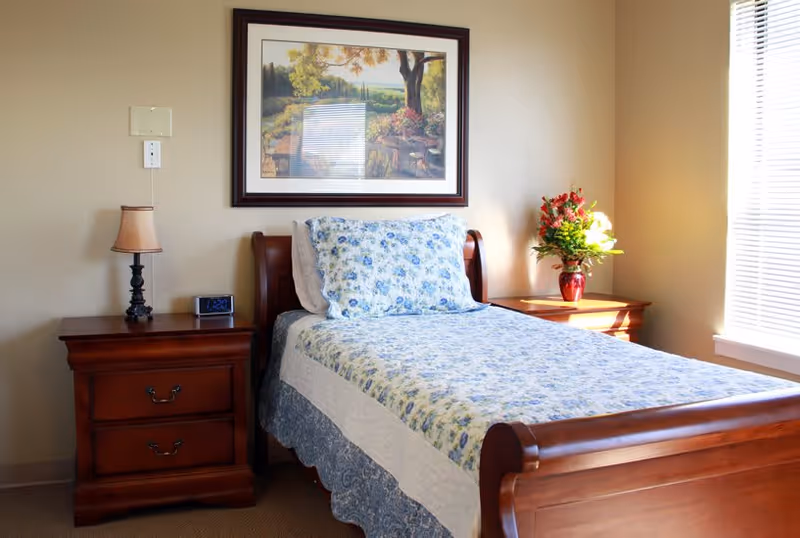 A neatly made single bed with a floral blue and white bedspread and matching pillow in a well-lit room. The bed has a wooden frame and is flanked by two wooden nightstands. One nightstand has a small lamp and a digital clock, while the other has a vase with a colorful flower arrangement. A framed landscape painting hangs on the beige wall above the bed, and sunlight streams in through a window with blinds on the right side.