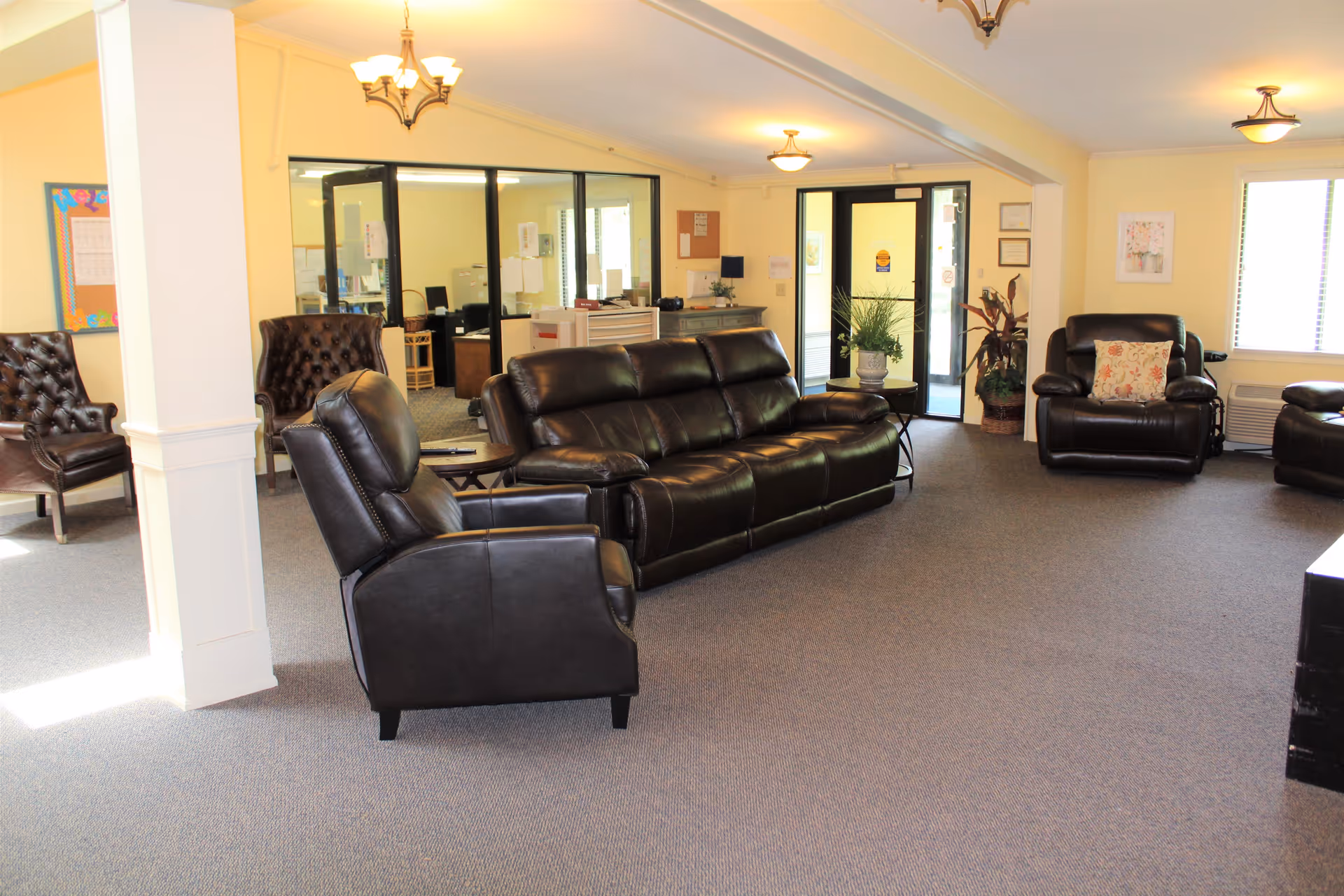 A spacious living room area with several dark brown leather recliners and a matching sofa arranged on a carpeted floor. The room has light yellow walls, multiple ceiling light fixtures, and large windows allowing natural light. There is a glass partition separating the living area from an office space in the background. Potted plants and framed artwork decorate the room.