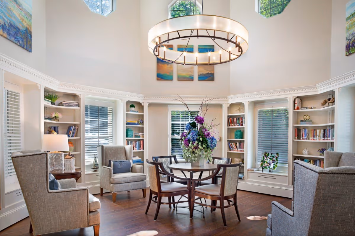 A bright and elegant living room with high ceilings, featuring a round table with four chairs in the center, a large floral arrangement on the table, and four upholstered armchairs arranged around it. The room has built-in bookshelves filled with books and decorative items, three windows with blinds, and colorful paintings on the walls. A large circular chandelier hangs from the ceiling.
