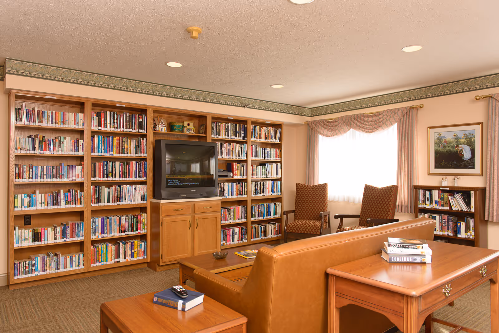A cozy living room area in Sanders Glen Assisted Living Center featuring a large wooden bookshelf filled with books, a television set in the center of the bookshelf, two patterned armchairs near a window with curtains, a leather sofa, and wooden tables with books and a remote control on them. The room has beige walls and carpeted floor.
