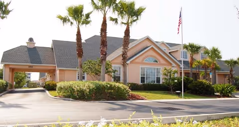 Peach-colored single-story senior living building front with a covered entrance, palm trees, landscaped shrubs, and an American flag.