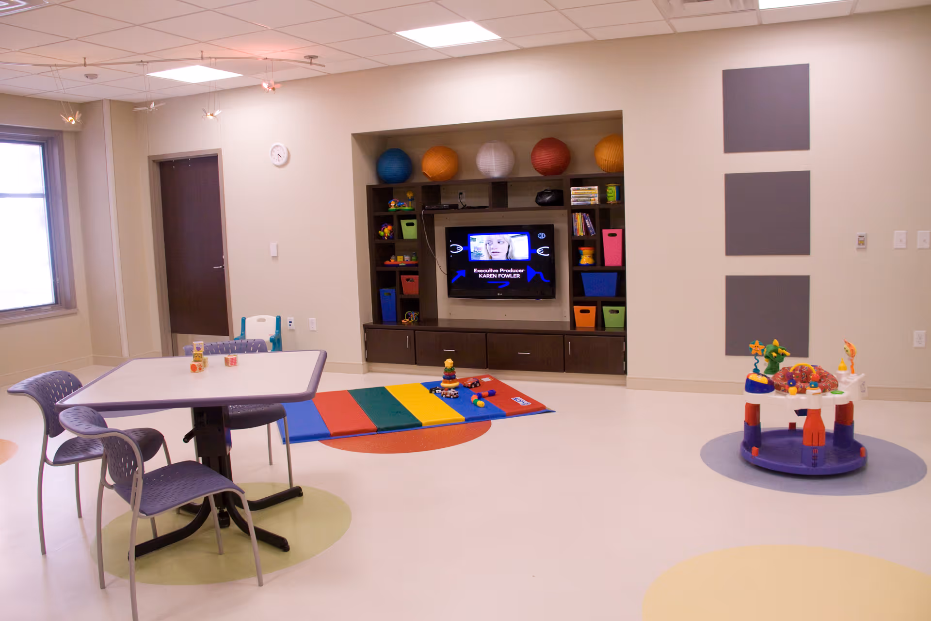 A bright, clean room with a table and chairs on the left, a colorful play mat with toys in the center, and a children's activity center on the right. A built-in shelving unit with a TV and colorful storage bins is set into the far wall. The room has a window, a door, and neutral-colored walls with three gray panels on the right wall.