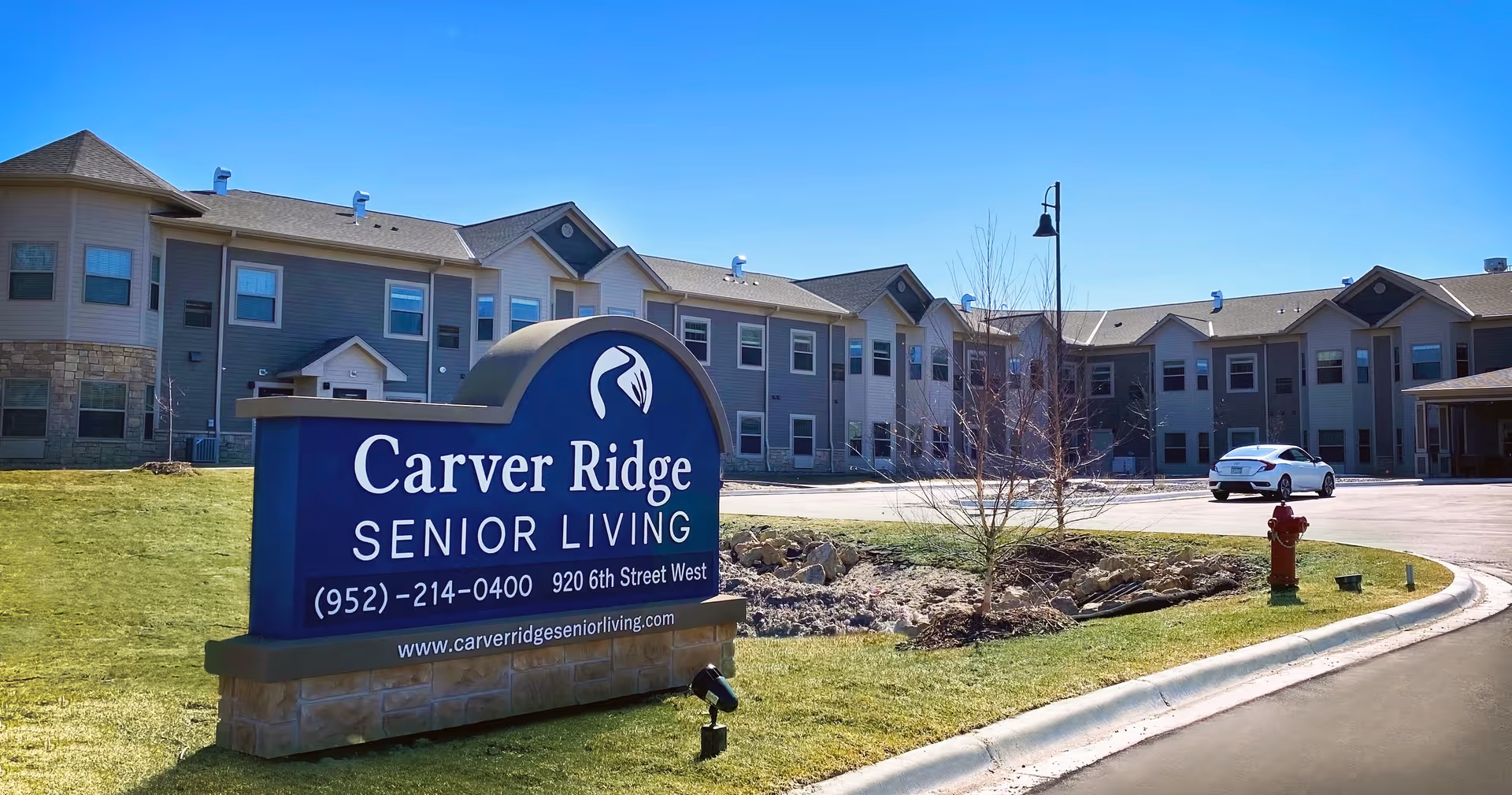 Exterior view of Carver Ridge Senior Living facility on a sunny day, showing a large two-story building with multiple windows, a parking area with a white car, a fire hydrant, and a prominent blue sign displaying the facility's name, phone number, address, and website.