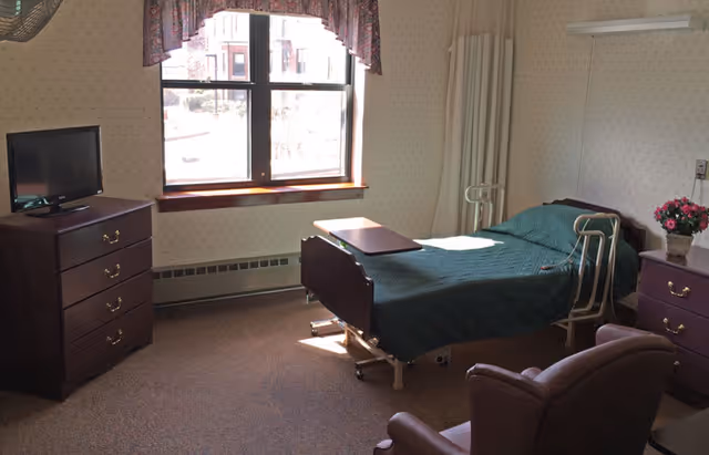 A senior living facility bedroom with a hospital-style bed covered in a green blanket, a small overbed table, a wooden dresser with a TV on top, a window with floral curtains, a purple armchair, and a nightstand with a flower pot.