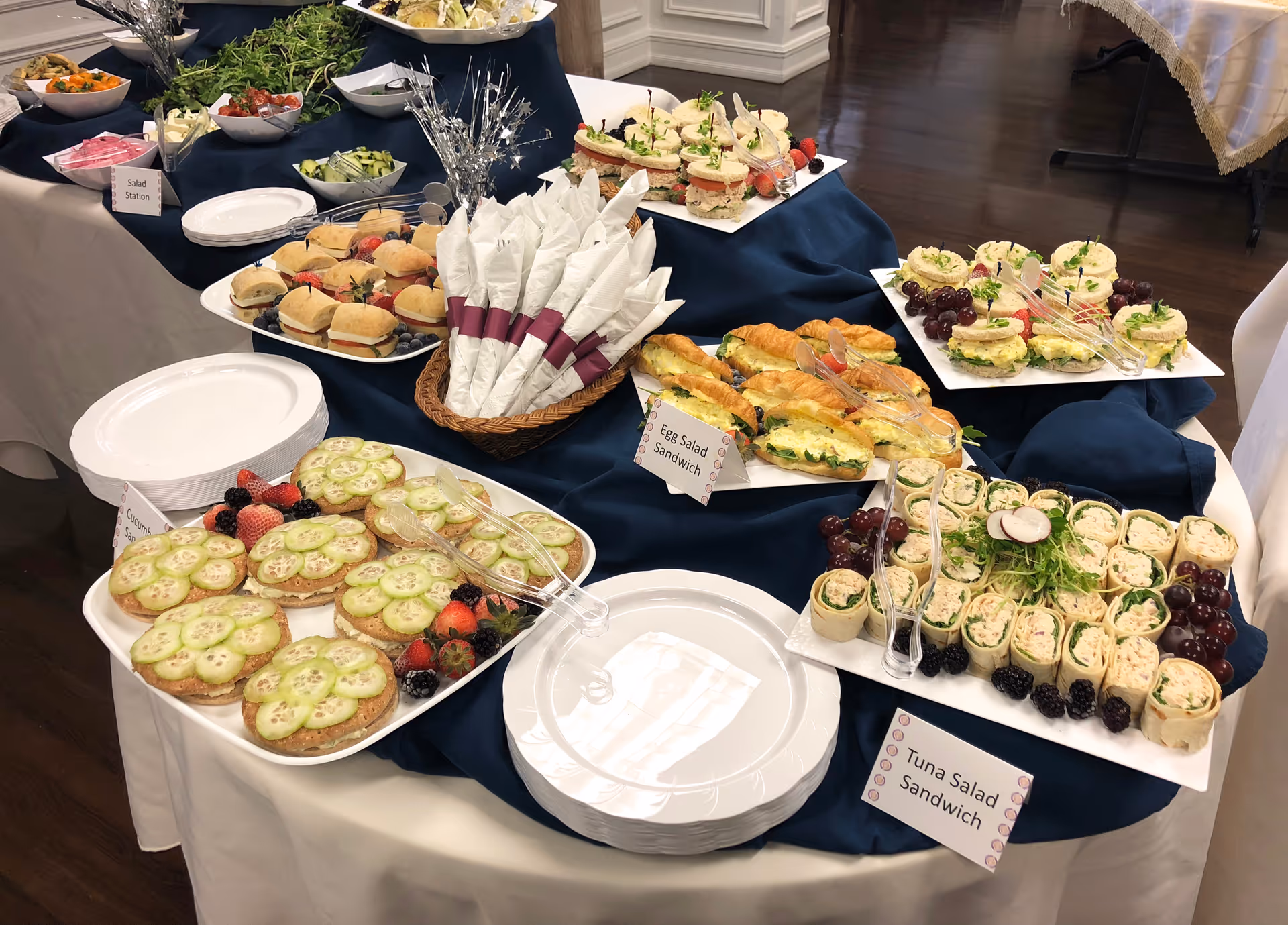 Buffet table with a variety of sandwiches and wraps including cucumber sandwiches, egg salad sandwiches, and tuna salad sandwiches, along with fresh berries and plates stacked for serving. The table is covered with a white tablecloth and a dark blue overlay.