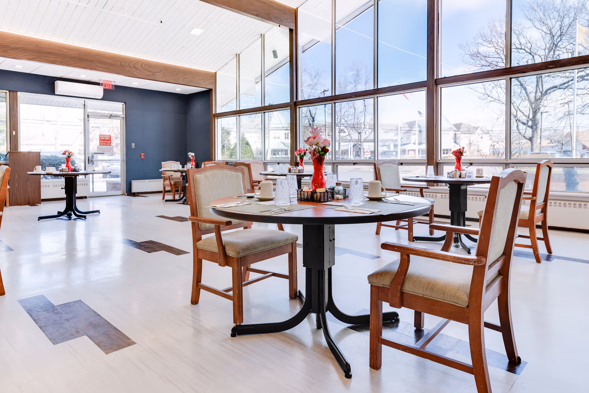 Bright dining room with large floor-to-ceiling windows letting in natural light. Several round tables are set with placemats, cups, glasses, and small flower vases. Wooden chairs with cushioned seats surround the tables. The room has light-colored flooring with darker wood inlays and a dark accent wall near the entrance door.