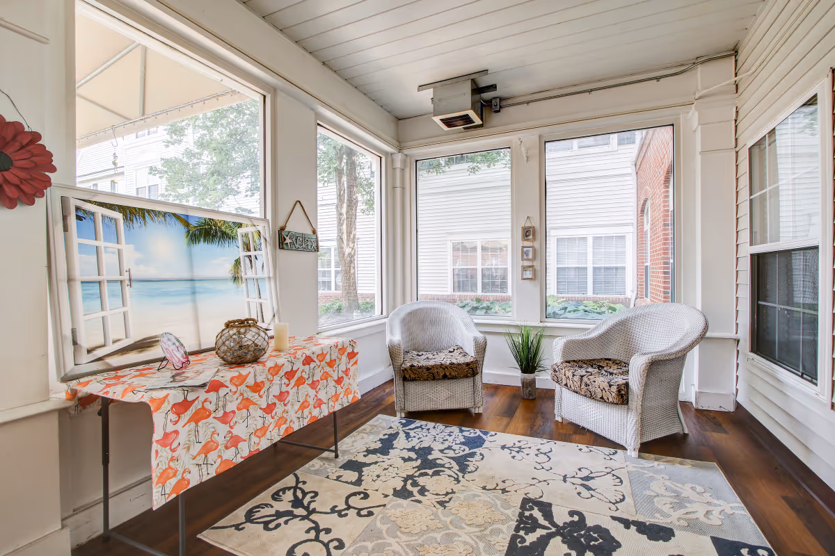 Bright enclosed sunroom with two wicker chairs, a patterned rug, and a table covered with a flamingo-print cloth beside large windows.