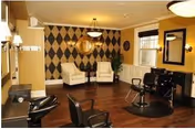 A well-lit interior salon/lounge with two white armchairs in front of a diamond-patterned accent wall, salon styling chairs, and shampoo stations on dark wood floors.
