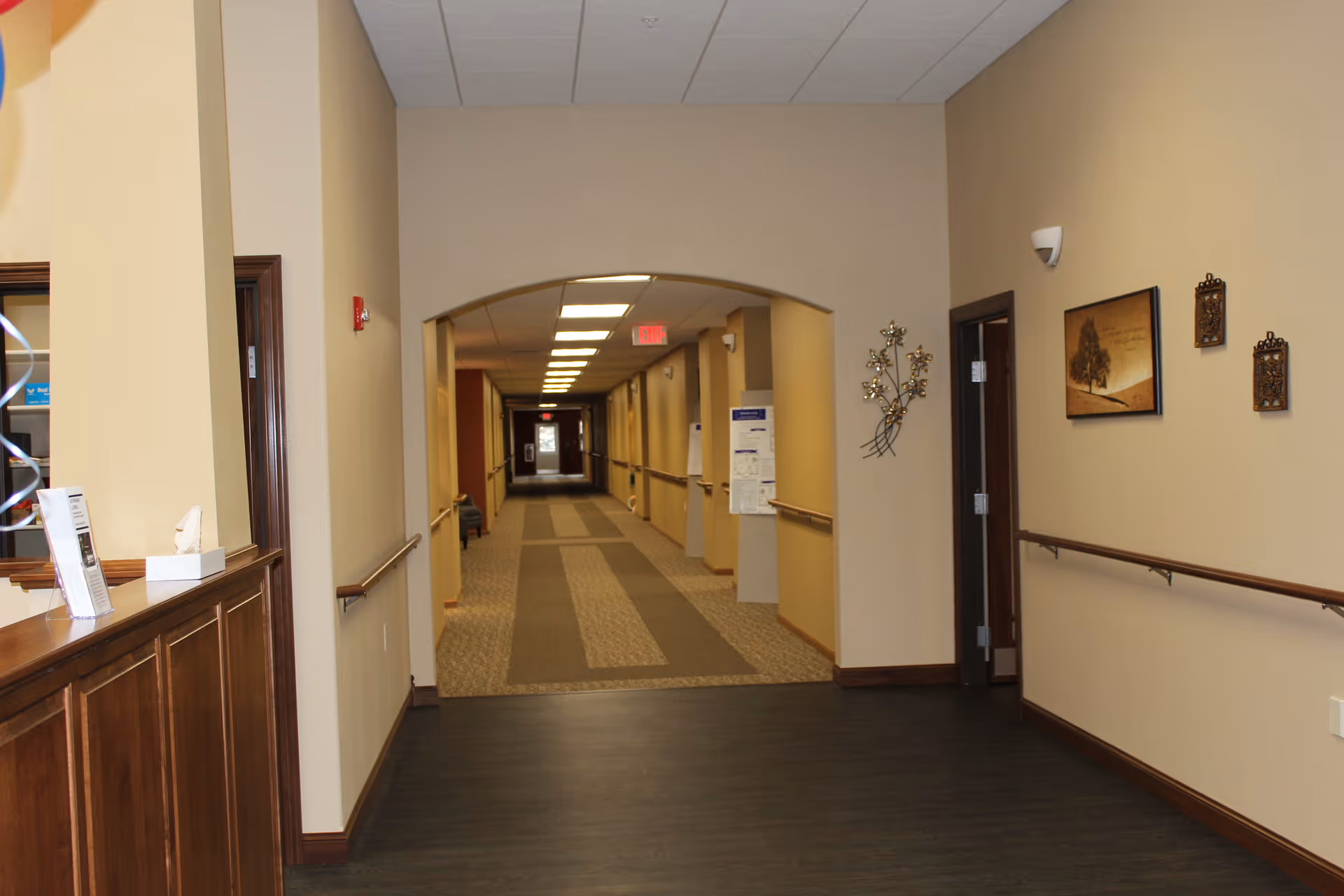 Interior hallway of a senior living facility with beige walls, wooden handrails on both sides, and a carpeted walkway. There is a wooden reception counter on the left side with some papers and a tissue box. The hallway extends into the distance with overhead fluorescent lighting and a red exit sign visible at the far end. Wall decorations include framed artwork and metal wall art on the right side.