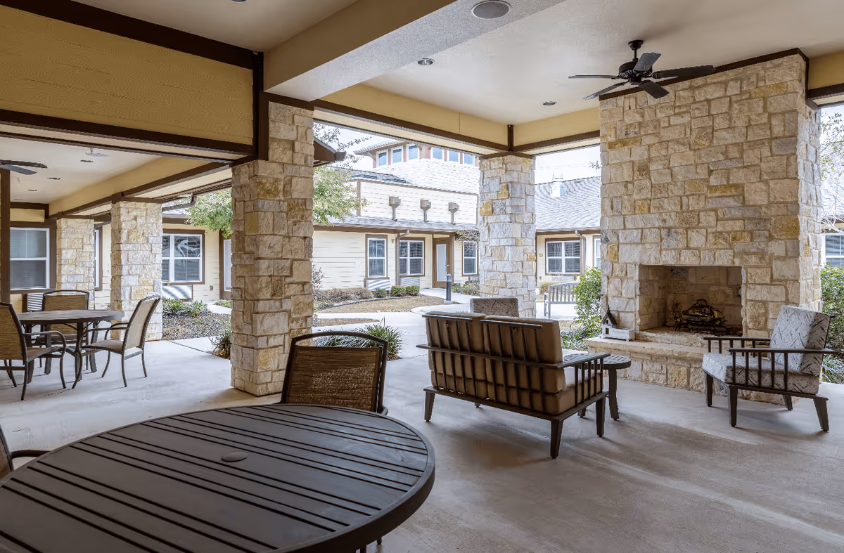 Covered outdoor patio area with stone pillars and a stone fireplace. The space includes cushioned seating, chairs, and tables, with ceiling fans overhead. Surrounding the patio are windows and doors leading to other parts of the facility.