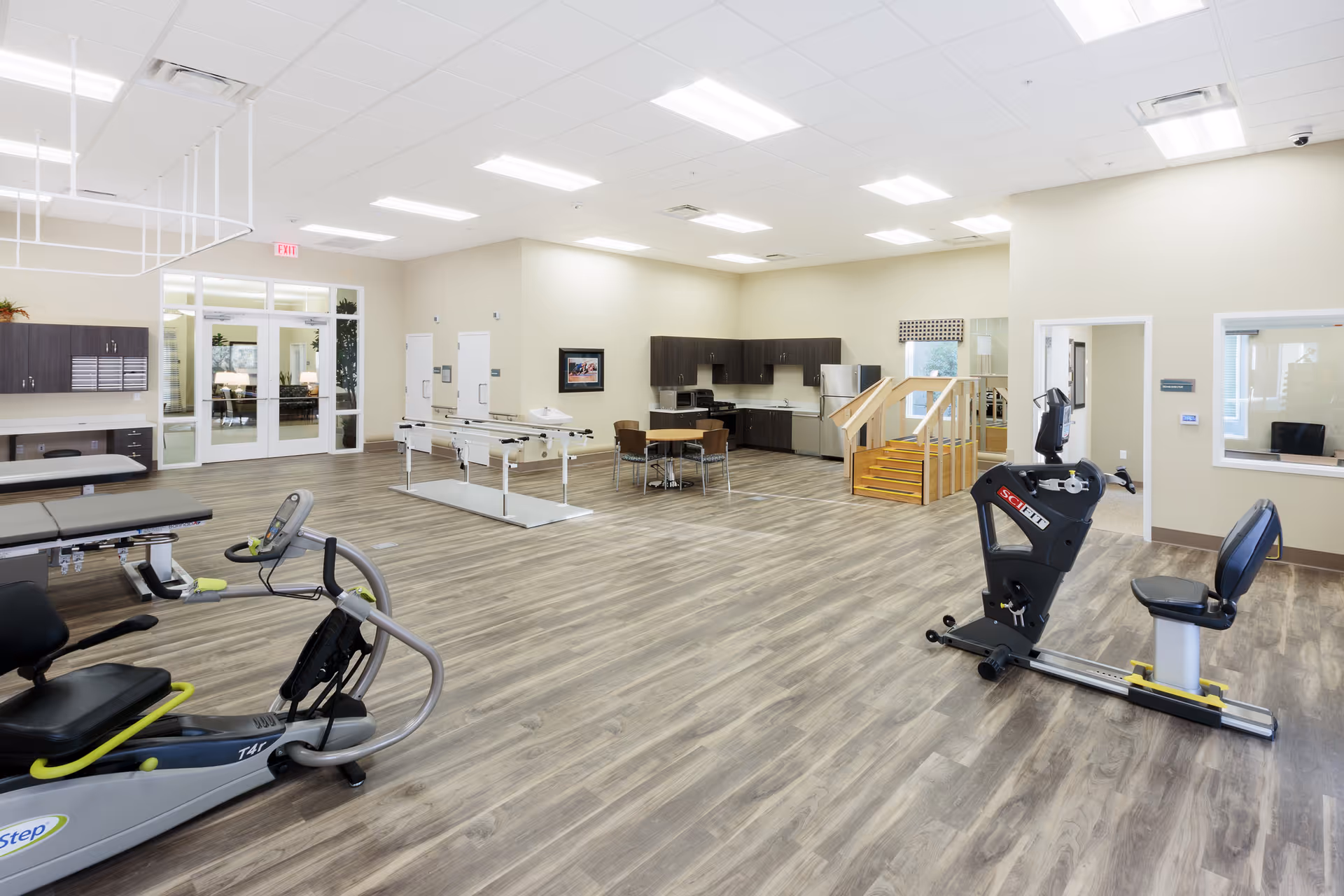 A spacious rehabilitation or physical therapy room with exercise equipment including a recumbent bike and parallel bars. The room has wood-look flooring, beige walls, and bright overhead lighting. There is a kitchenette area with cabinets, a table with chairs, and a small set of wooden stairs with handrails. Large glass doors and windows are visible in the background.
