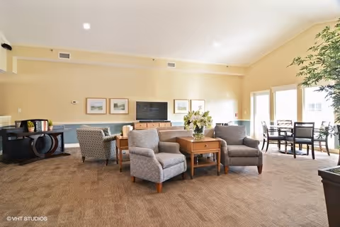 A spacious and well-lit living room area with beige walls and carpeted floor. The room features several gray upholstered armchairs arranged around wooden side tables with a vase of flowers. A flat-screen TV is mounted on the wall above a wooden cabinet. There are framed pictures on the wall and a dining table with chairs near large windows letting in natural light. A large potted plant is visible on the right side of the room.