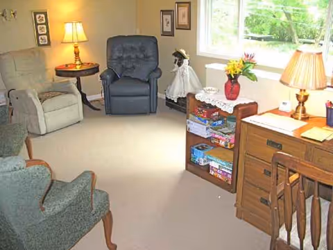 Cozy senior living room with recliners, a desk, bookshelf, and a window overlooking greenery.