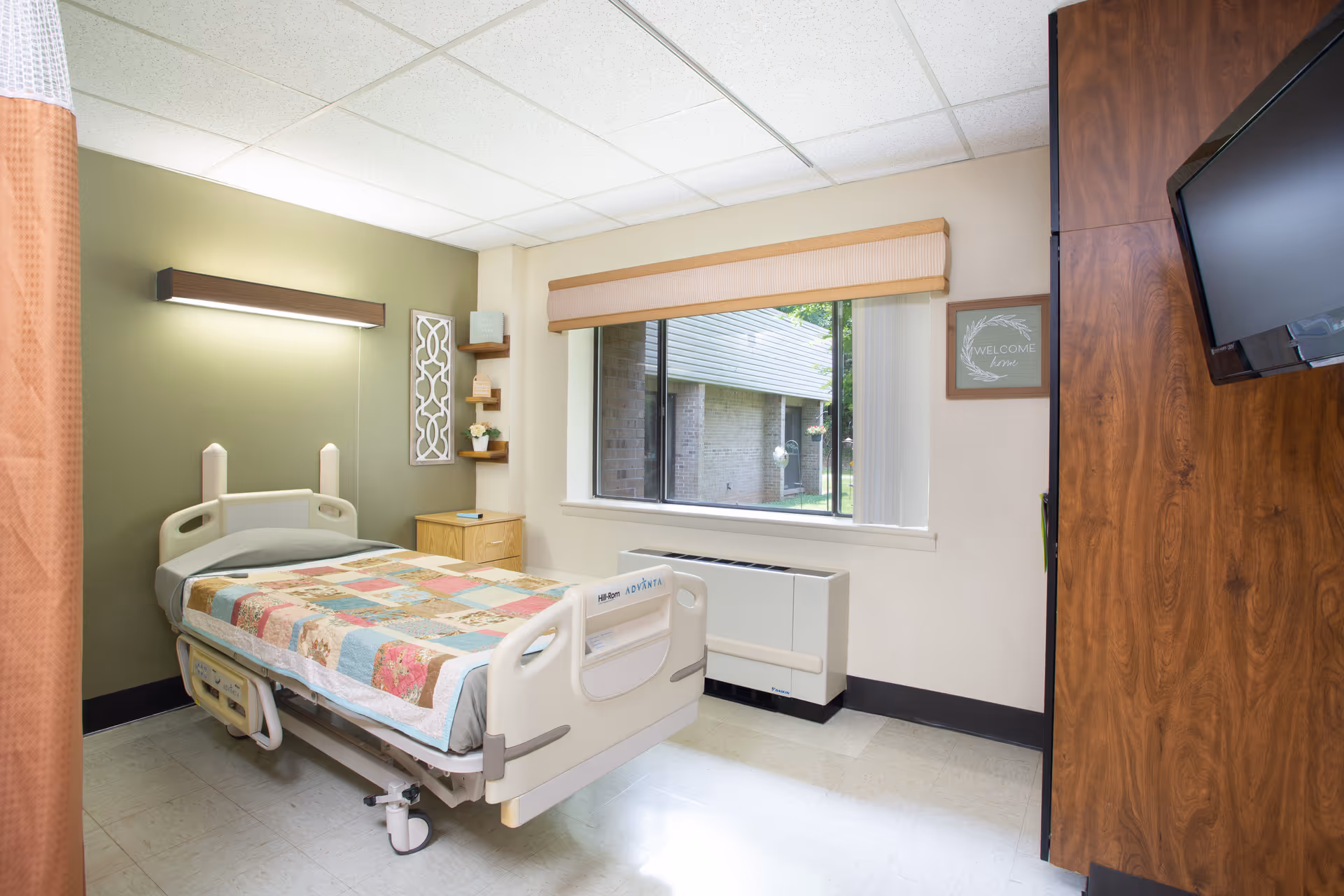 A small, clean patient room with a single hospital bed covered with a colorful patchwork quilt. The room has a green accent wall with a light fixture above the bed, a wooden nightstand with small shelves holding decorative items, a large window with a beige valance, and a wall-mounted TV on the right side.