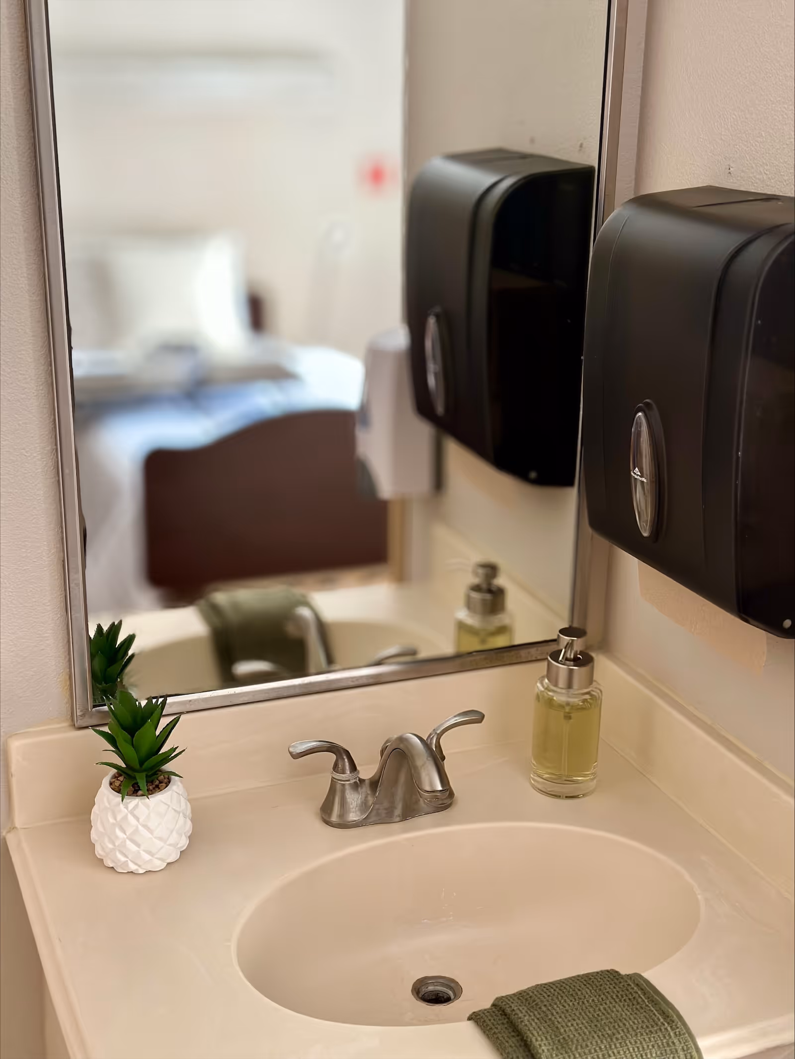 A bathroom vanity with a sink, faucet, liquid soap dispenser, wall-mounted paper towel dispenser, small potted plant, and a mirror reflecting a bed.