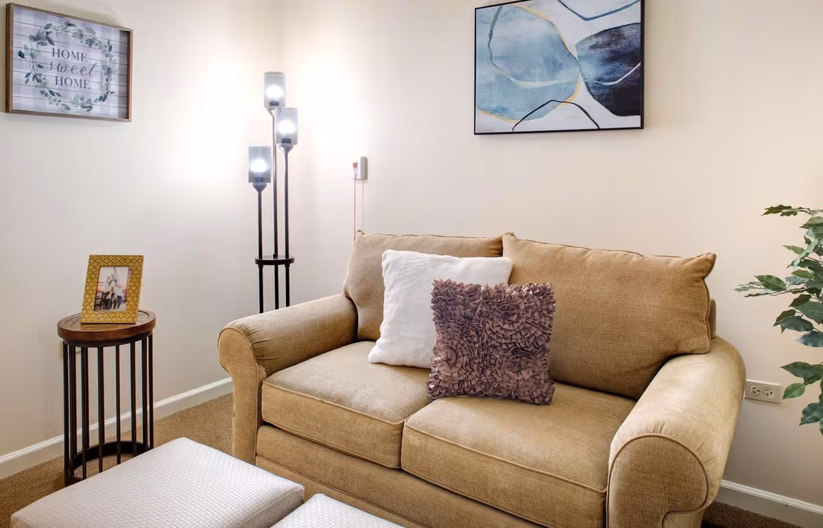 A cozy living room area with a beige couch adorned with two decorative pillows, one white and one textured purple. Next to the couch is a small round wooden side table with a framed photo. A modern floor lamp with three lights stands behind the table. On the wall above the couch is an abstract blue and white painting, and to the left is a framed sign that reads 'Home Sweet Home'. A green plant is partially visible on the right side.