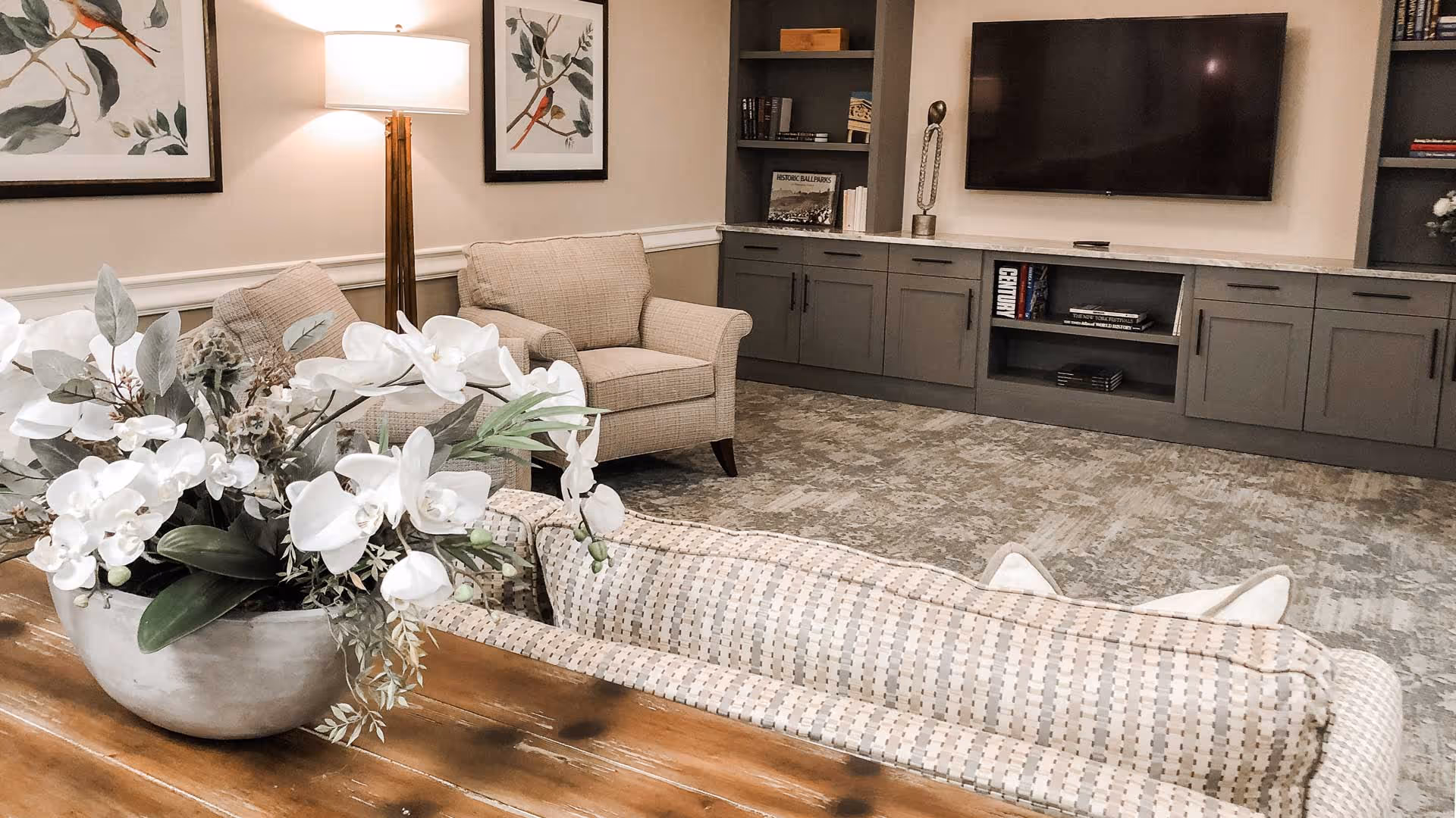 A cozy living room area with beige armchairs, a patterned sofa, a wooden table with a large white floral arrangement, a floor lamp, framed bird artwork on the wall, and a built-in gray entertainment center with a flat-screen TV and shelves holding books and decorative items.