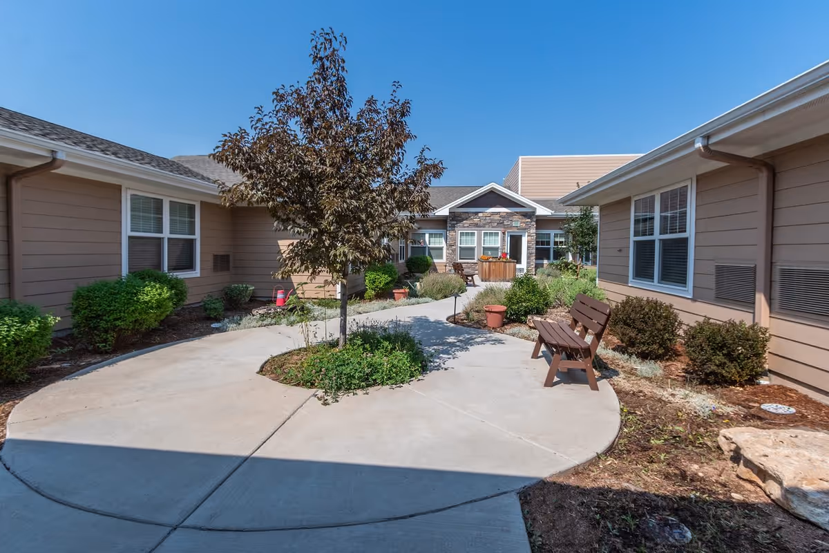Outdoor courtyard area at Willowbrook Place with a concrete pathway curving around a small tree and garden bed. There are benches along the pathway and shrubs planted near the beige buildings under a clear blue sky.