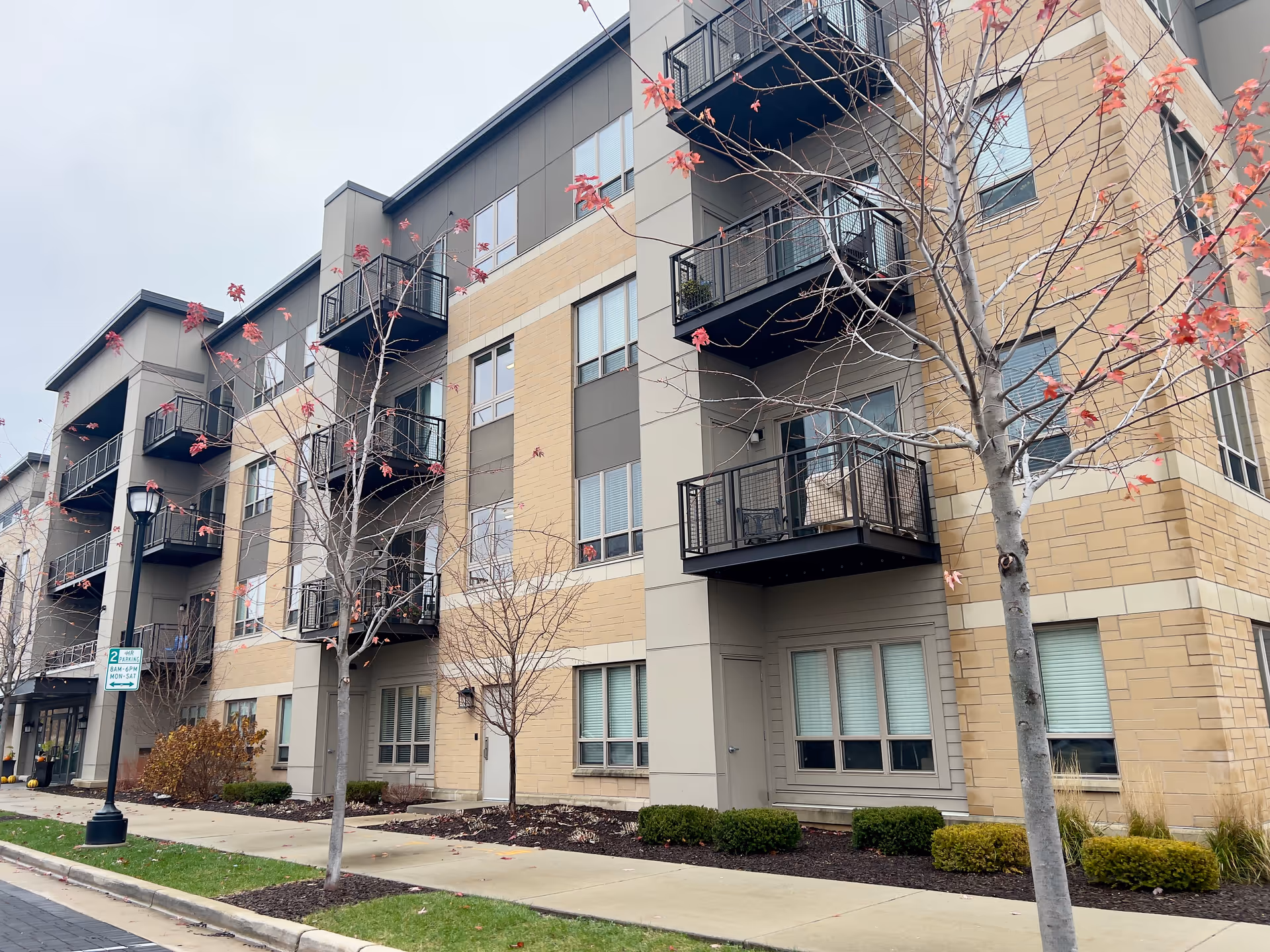 Front exterior of a multi-story residential building with balconies, trees, and a sidewalk.