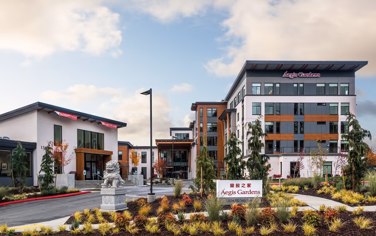 Exterior view of Aegis Gardens Newcastle senior living facility showing modern multi-story buildings with large windows, landscaped gardens with shrubs and trees, a stone lion statue, and a sign with the facility name in English and Chinese.