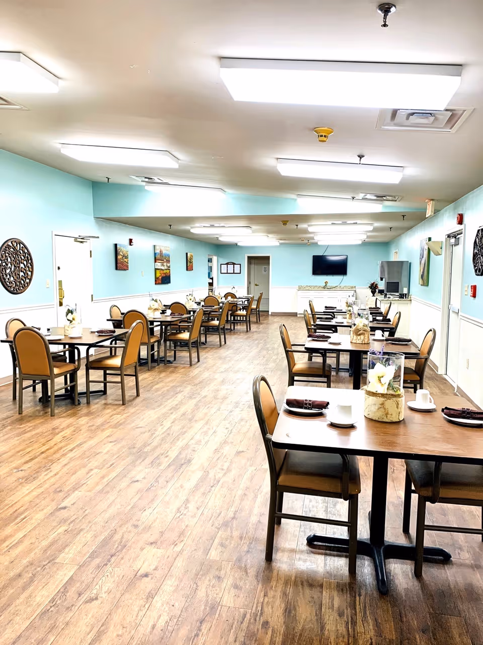A spacious dining room with multiple tables and chairs arranged neatly. Each table has place settings including cups, plates, and napkins, along with decorative centerpieces. The room has light blue walls with white wainscoting, wood flooring, and bright overhead lighting. There are paintings and decorative wall hangings on the walls, and a television mounted on the far wall.