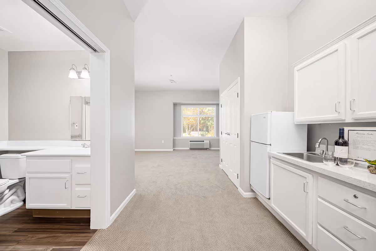 Interior view of a senior living facility apartment at Ivy Park at Tustin showing a small kitchen area with white cabinets, a sink, and a refrigerator on the right. To the left is a bathroom with a toilet and vanity visible. The space opens into a carpeted living area with a large window and an air conditioning unit below it.