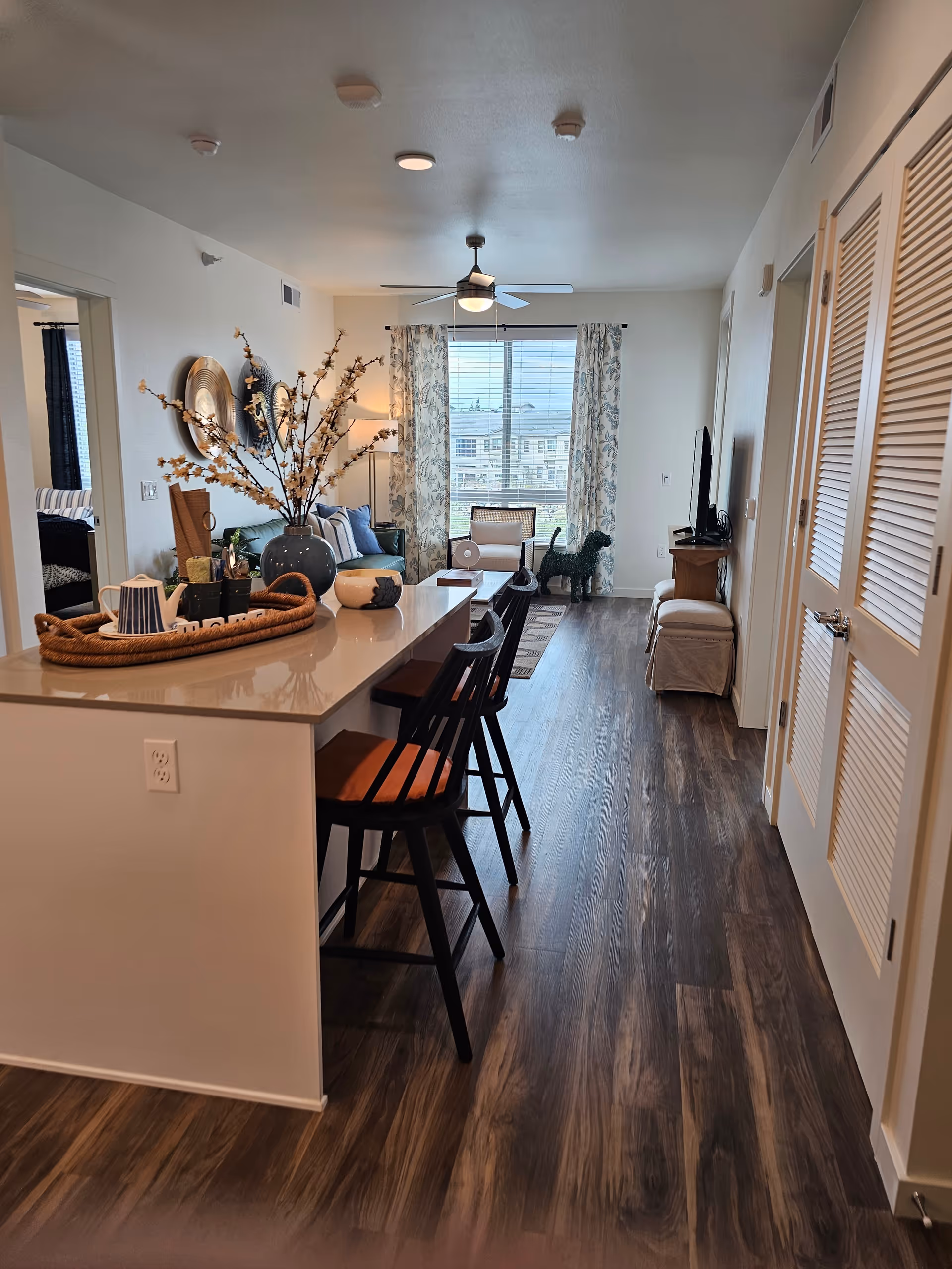 Interior view of a modern apartment with a kitchen island and three bar stools in the foreground. The kitchen island has decorative items including a vase with flowers and a tray with a teapot. Beyond the island is a living room area with a sofa, armchair, coffee table, and a TV on a stand. A large window with patterned curtains lets in natural light. The floor is wood, and there are doors and closets visible along the right side.
