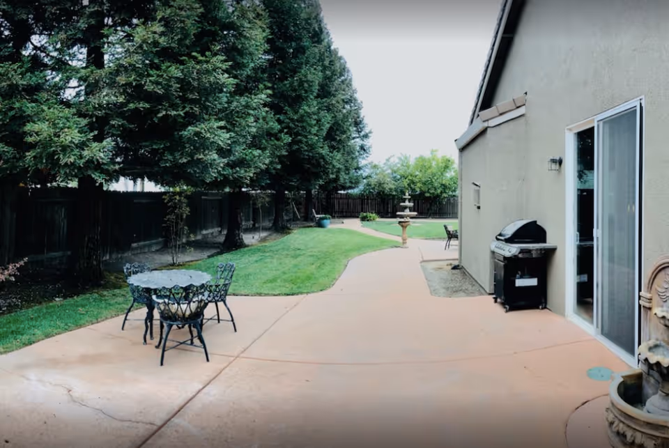 Patio area with a metal table and chairs, a barbecue grill by a sliding door, a paved walkway leading to a fountain, and trees along a fenced yard.