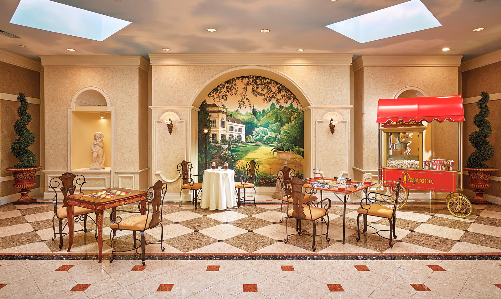 An elegant indoor seating area with decorative wrought iron chairs and tables on a checkered marble floor. The back wall features a large mural of a garden scene with a house, framed by an archway. There is a red popcorn cart on the right side and two spiral topiary plants in large pots on either side of the room. The ceiling has recessed lighting and two skylights.
