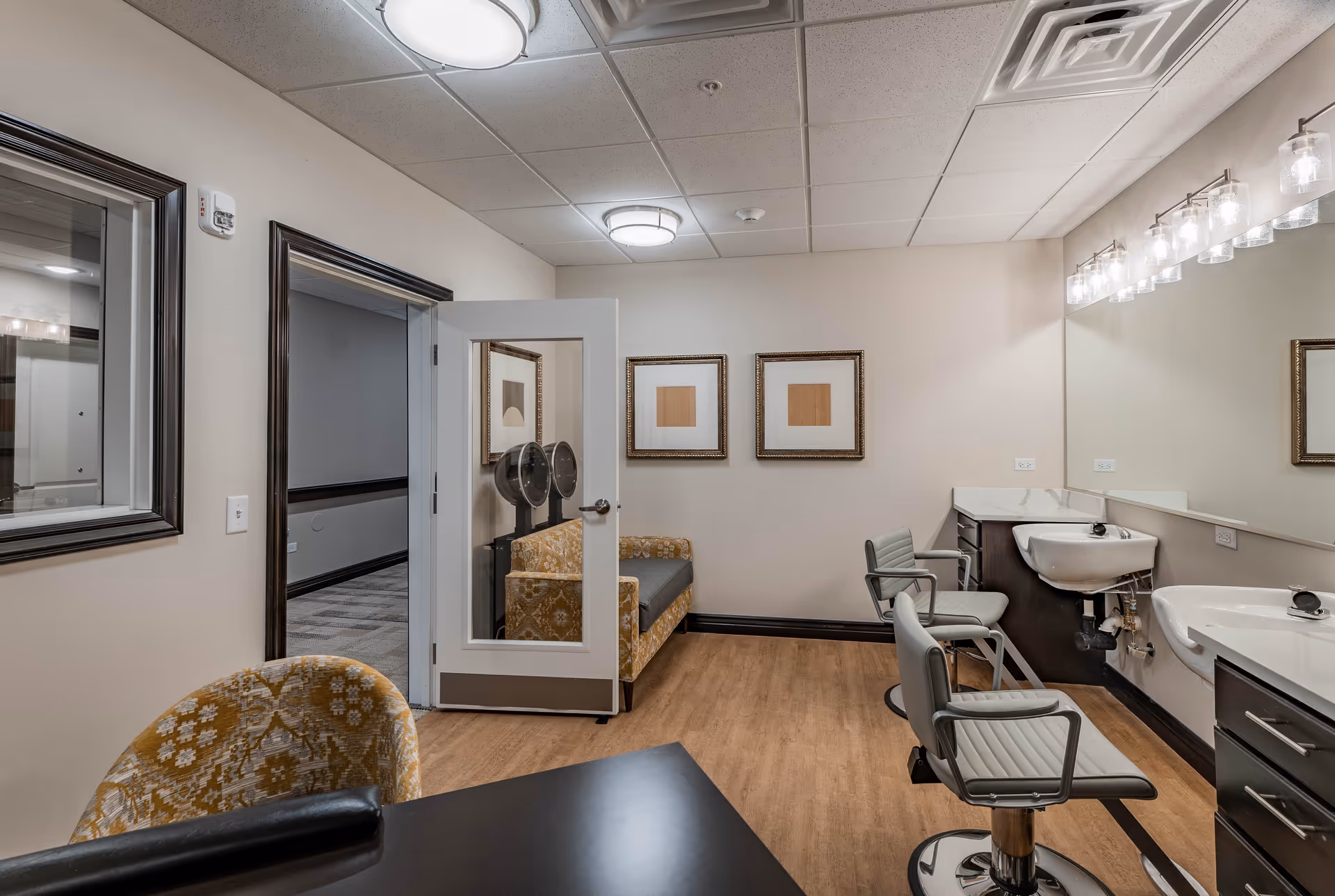 Interior view of a senior living facility hair salon area with two salon chairs facing sinks and a large mirror. There is a small table with a patterned chair in the foreground and a yellow patterned couch against the far wall. The room has light-colored walls, wood flooring, and ceiling lights.