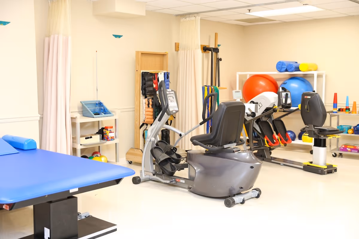 A physical therapy or rehabilitation room with exercise equipment including recumbent bikes, therapy balls, and shelves holding various therapy tools and supplies. There is also a blue therapy table and privacy curtains in the background.