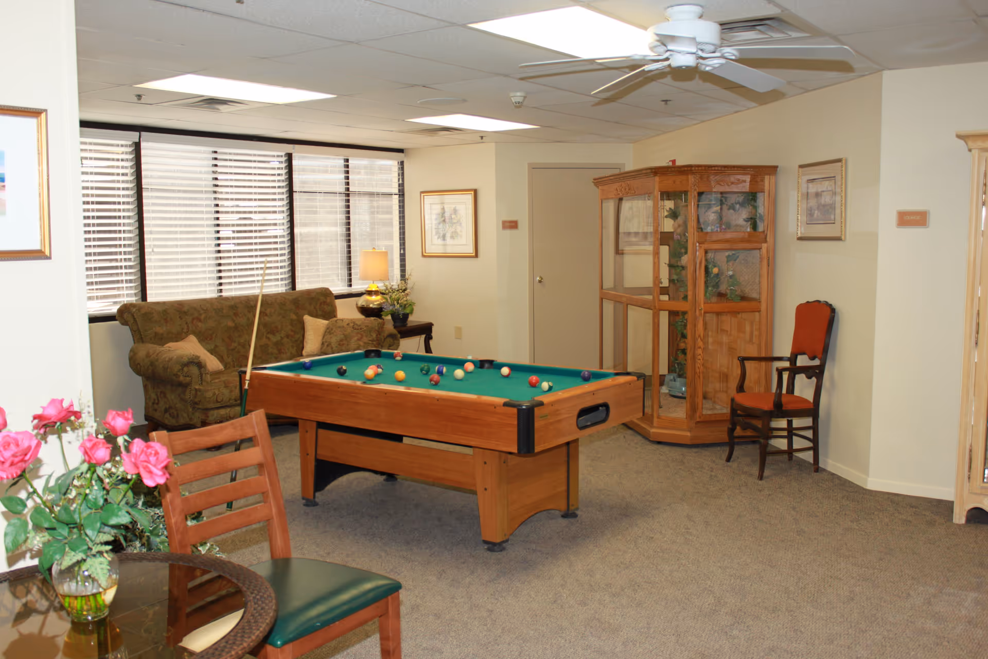 A cozy lounge area with a pool table in the center, a floral patterned couch with pillows near a window with blinds, a wooden display cabinet with plants inside, a wooden chair with red upholstery, a small round glass table with a vase of pink roses, and a ceiling fan above.